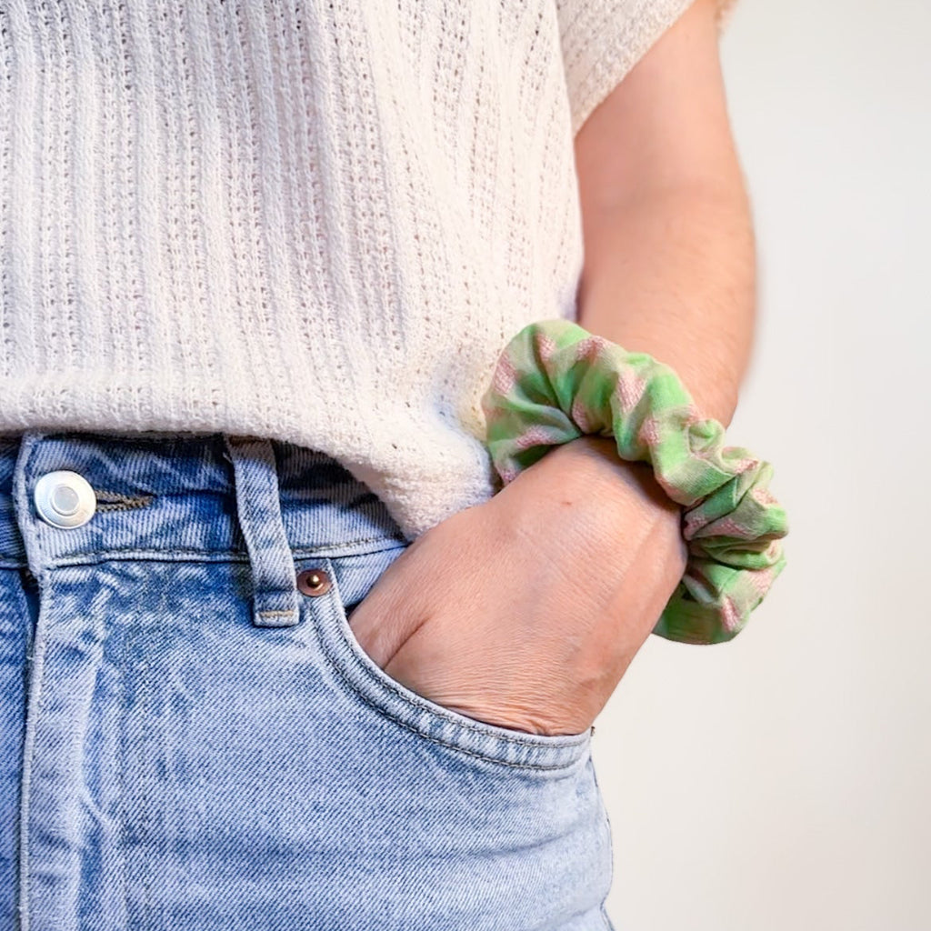 Gros plan sur un chouchou artisanal Jolie Myrtille en tissu à petits carreaux vichy (gingham) vert pastel et rose, porté au poignet d'une personne avec une main dans la poche de son jean bleu clair.