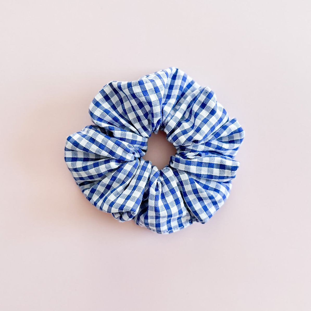 Gros chouchou cheveux en motif vichy bleu et blanc de fabrication artisanale en Provence, accessoire de coiffure femme tendance signé Jolie Myrtille, présenté à plat sur un fond rose pastel.
