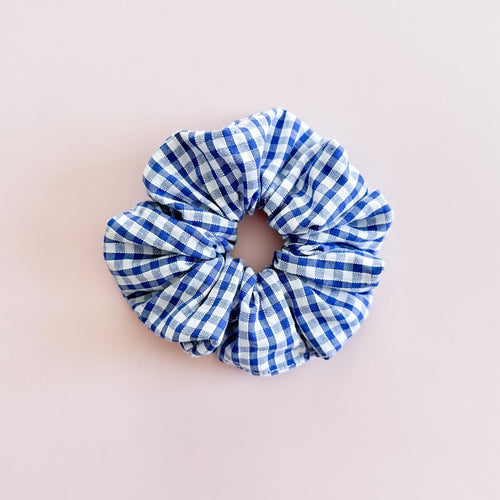 Gros chouchou cheveux en motif vichy bleu et blanc de fabrication artisanale en Provence, accessoire de coiffure femme tendance signé Jolie Myrtille, présenté à plat sur un fond rose pastel.