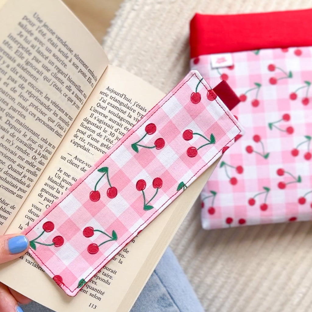 Marque-page en tissu cousu main motif vichy rose et cerises, posé sur un livre avec sa pochette de lecture assortie en arrière plan.