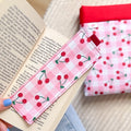 Marque-page en tissu cousu main motif vichy rose et cerises, posé sur un livre avec sa pochette de lecture assortie en arrière plan.
