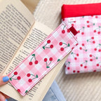 Marque-page en tissu cousu main motif vichy rose et cerises, posé sur un livre avec sa pochette de lecture assortie en arrière plan.