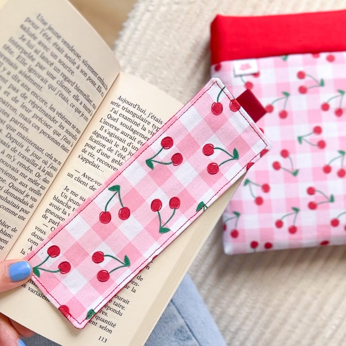 Marque-page en tissu cousu main motif vichy rose et cerises, posé sur un livre avec sa pochette de lecture assortie en arrière plan.