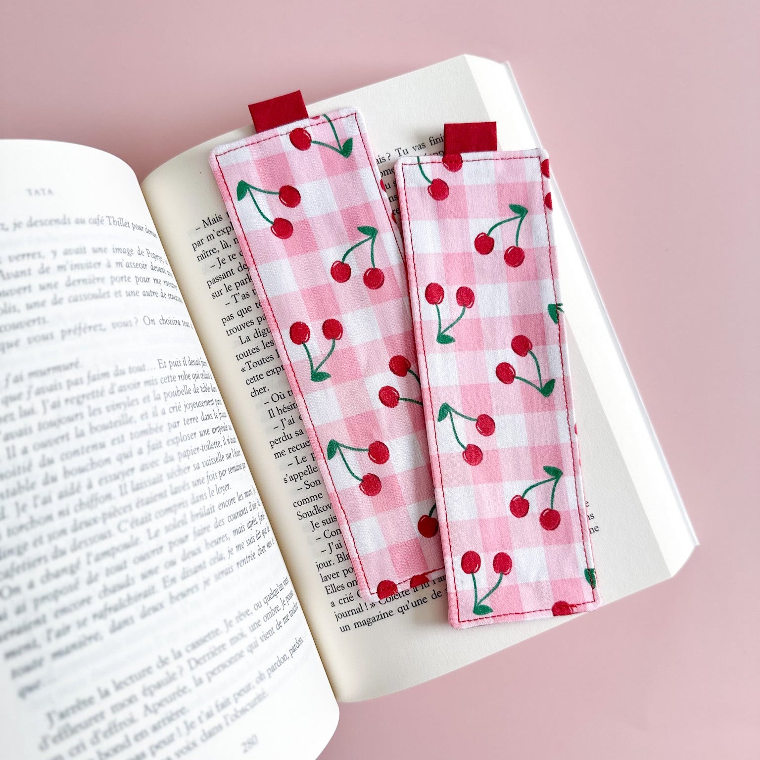 Deux jolis marque-pages en tissu fait main avec un motif vichy rose et des cerises rouges, posées sur les pages d'un roman.
