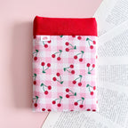 Vue de face d'une pochette de protection pour livre en coton vichy rose imprimé cerises avec une bordure en vichy rouge.