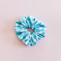 Photo de produit à plat du chouchou artisanal Marina, accessoire de coiffure pour femme au motif à carreaux Vichy bleu ciel et blanc, présenté sur un fond uni rose clair. Idéal comme idée de cadeau.