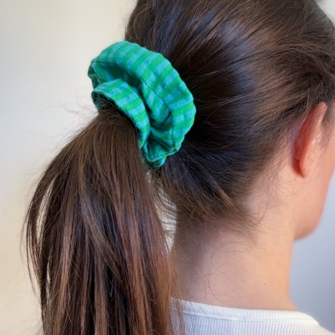 Femme brune aux longs cheveux lisses avec une queue-de-cheval attachée par le chouchou Siena à rayures vertes et bleu turquoise en gaze de coton, accessoire de fabrication artisanale.