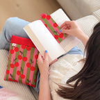 Une lectrice tenant son roman ouvert avec le marque page en tissu rayé et cerises, accompagnée de sa pochette à livre assortie sur les genoux.