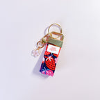 Bijou de sac et porte-clés Charlotte en tissu motif fraises et fleurs posé à plat sur fond rose, accessoire fait main avec sa boucle dorée et sa breloque fleur.