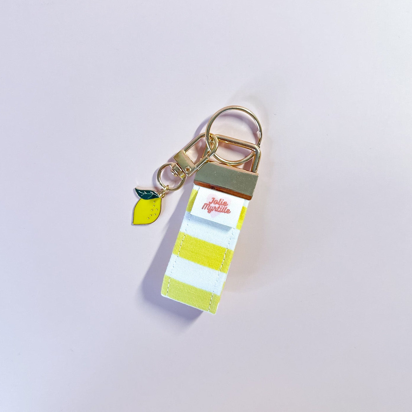 Bijou de sac et porte-clés Limoncello en tissu à rayures posé à plat sur fond rose, accessoire fait main avec sa boucle dorée et sa breloque citron.