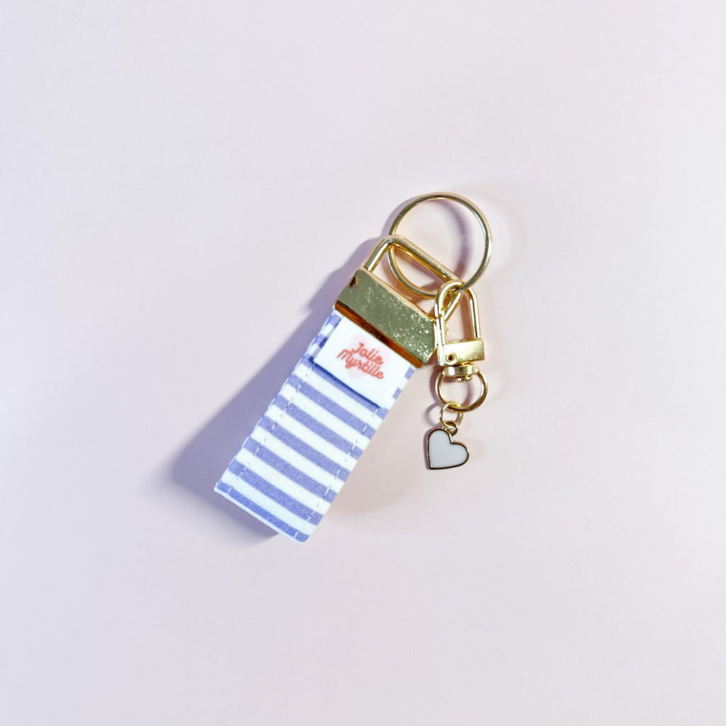Bijou de sac et porte-clés Anabelle en tissu à rayures posé à plat sur fond rose, accessoire fait main avec sa boucle dorée et sa breloque cœur.