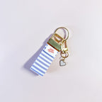 Bijou de sac et porte-clés Anabelle en tissu à rayures posé à plat sur fond rose, accessoire fait main avec sa boucle dorée et sa breloque cœur.