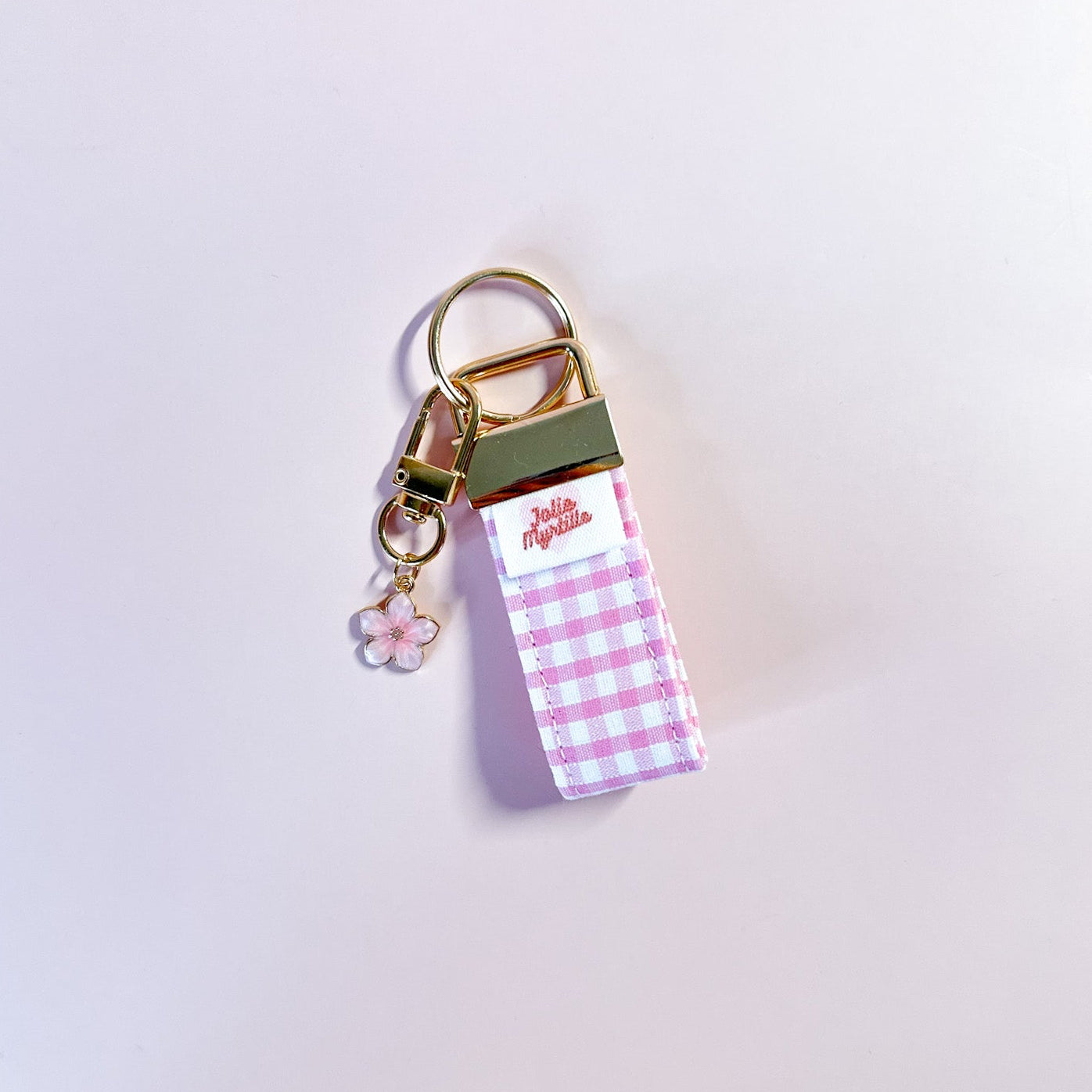 Bijou de sac et porte-clés Rosie en tissu vichy posé à plat sur fond rose, accessoire fait main avec sa boucle dorée et sa breloque fleur rose.