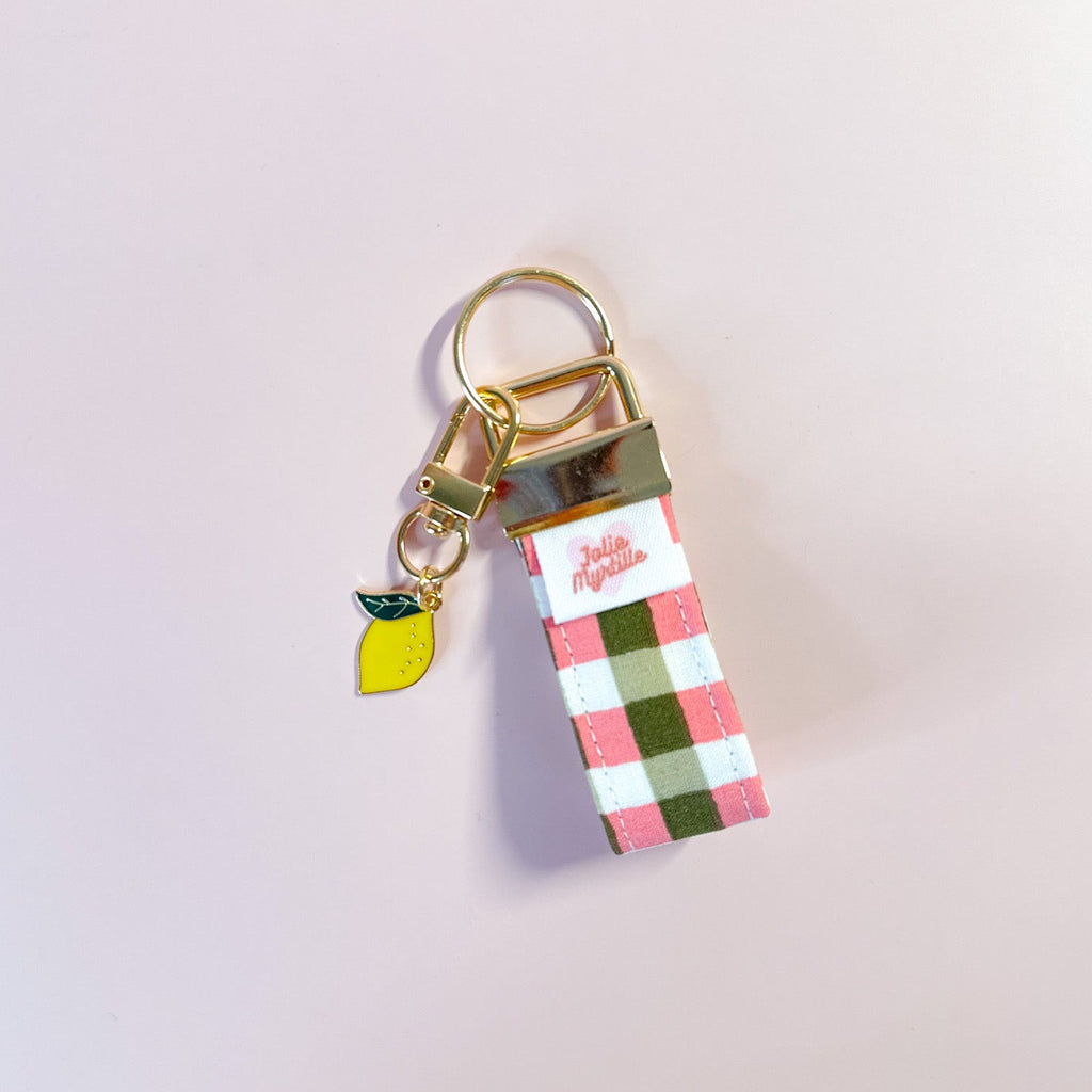 Bijou de sac et porte-clés Zita en tissu vichy posé à plat sur fond rose, accessoire fait main avec sa boucle dorée et sa breloque citron.