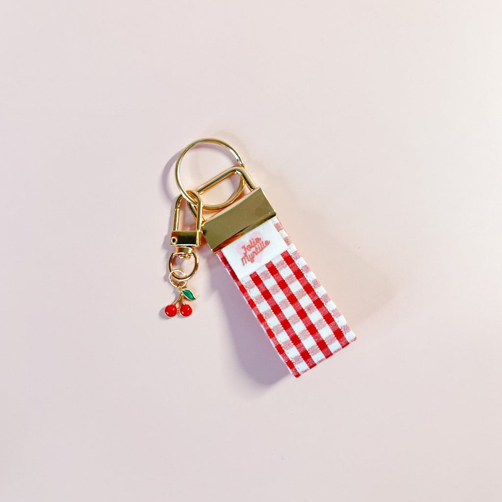 Bijou de sac et porte-clés Anna en tissu vichy posé à plat sur fond rose, accessoire fait main avec sa boucle dorée et sa breloque cerise.