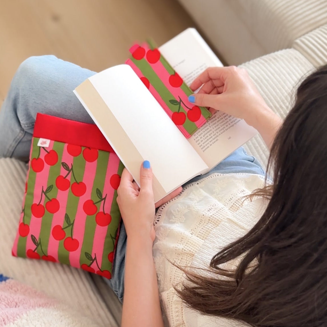 femme assise tenant un livre dans ses mains avec un marque pages en tissu cousu main et une pochette à livre imprimés cerise rouge à rayures rose et verte.
