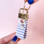 Main tenant le mini porte-clés Anabelle en tissu à rayures mauves et blanches, montrant le mousqueton doré et son petit charm en forme de cœur. Fabrication artisanale.
