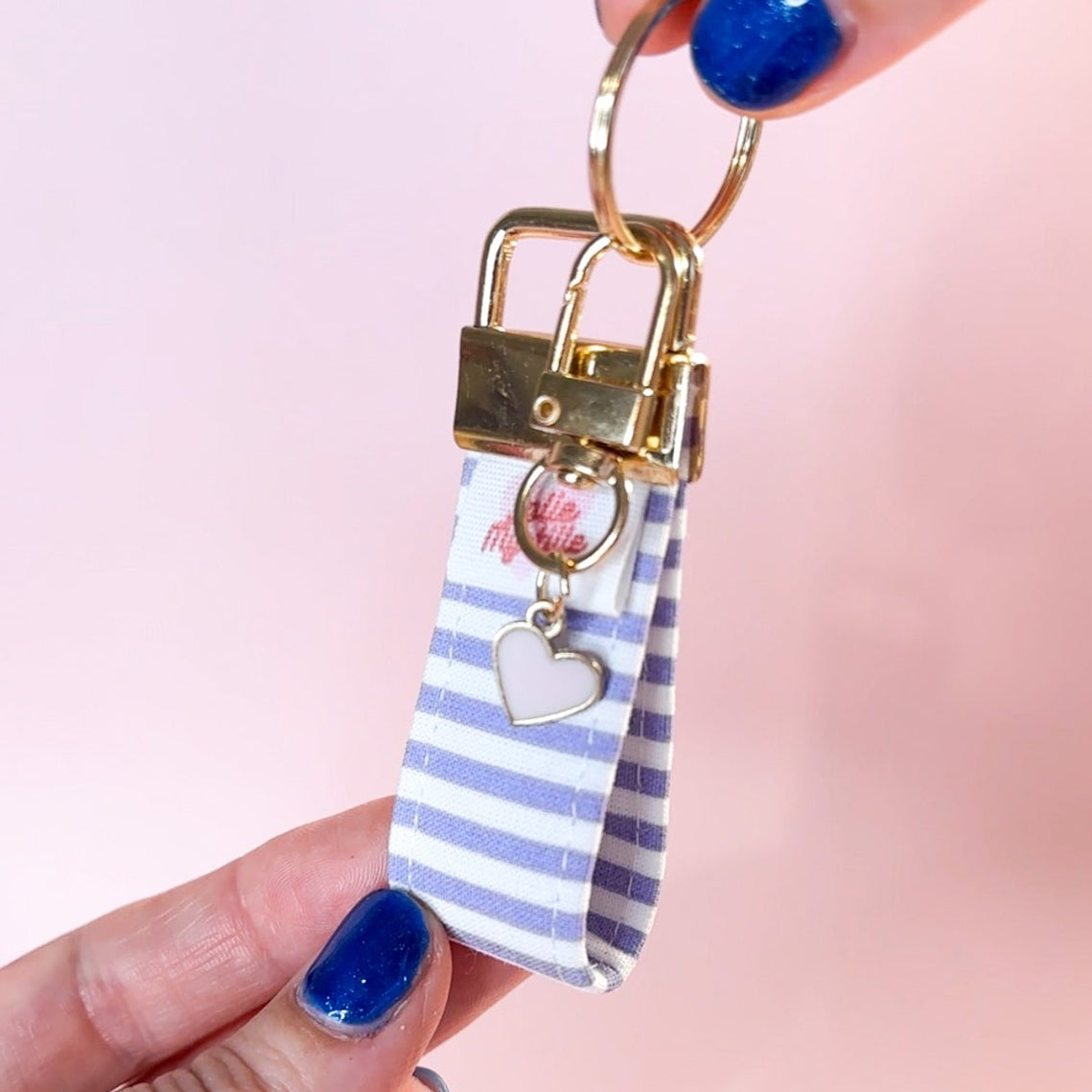 Main tenant le mini porte-clés Anabelle en tissu à rayures mauves et blanches, montrant le mousqueton doré et son petit charm en forme de cœur. Fabrication artisanale.