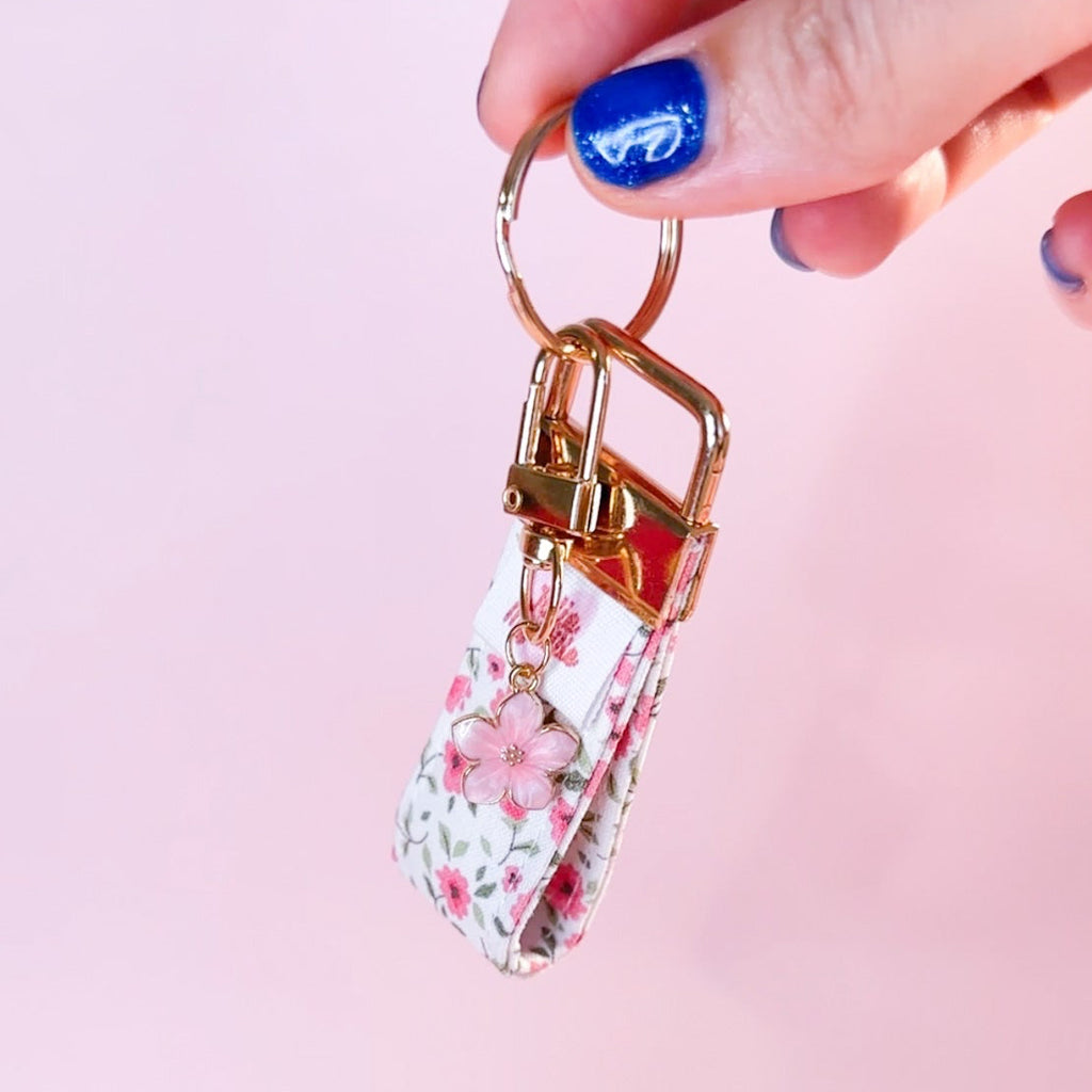 Main tenant le mini porte-clés Anita en tissu motif fleuri, montrant le mousqueton doré et son charm fleur rose. Produit fait main.
