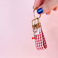 Main tenant le mini porte-clés Anna en tissu vichy rouge et blanc, montrant le mousqueton doré et son petit charm cerise. Produit fait main.