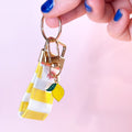 Main tenant le mini porte-clés Limoncello en tissu à rayures jaunes et blanches, montrant le mousqueton doré et son petit charm citron. Fabrication artisanale.