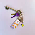 Porte-clés Nina en tissu vichy orange accroché à un trousseau de clés, utilisé comme un grigri de sac fruité et pratique au quotidien.