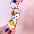Main tenant le mini porte-clés Nina en tissu vichy orange, rose et blanc, montrant le mousqueton doré et son petit charm citron. Produit fait main.