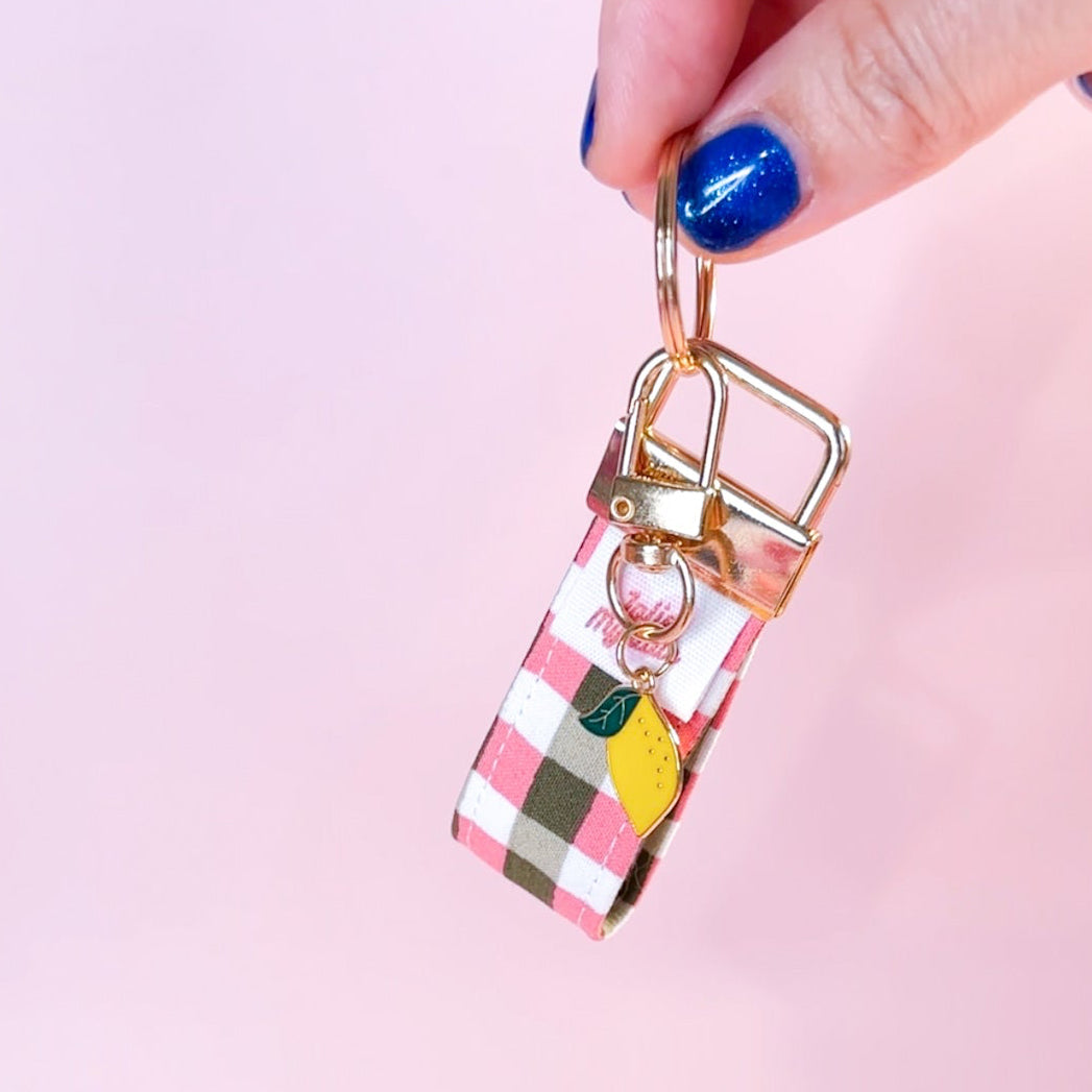 Main tenant le mini porte-clés Zita en tissu motif vichy rose et vert kaki, montrant le mousqueton doré et son petit charm citron. Fabrication artisanale.
