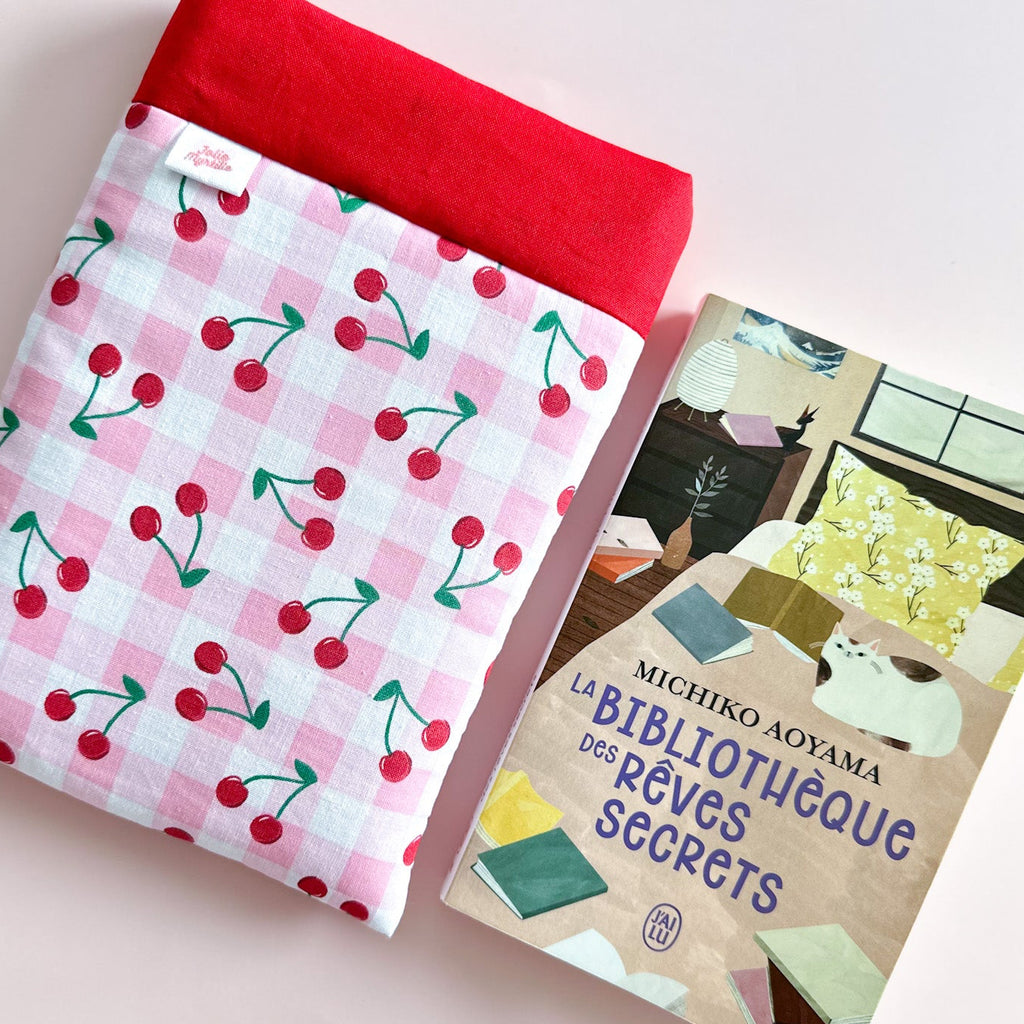 Pochette à livre Jolie Myrtille Fabric en tissu coton vichy rose et cerises posé à coté d'un roman pour illustrer le format de cette housse de protection artisanale.