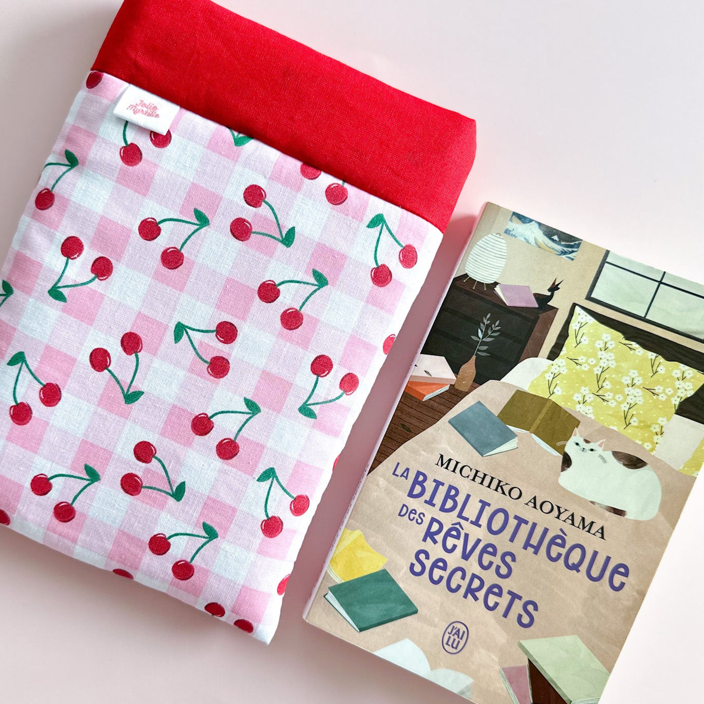 Pochette à livre Jolie Myrtille Fabric en tissu coton vichy rose et cerises posé à coté d'un roman pour illustrer le format de cette housse de protection artisanale.